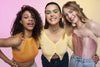 Three joyful friends showcasing vibrant Gen Z makeup styles and trendy hairstyles, embodying the concept of a diverse hair cycle with their unique hair textures and colors, standing against a pink background
